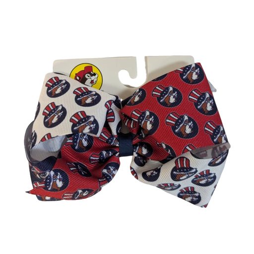 A patriotic Buc-ee's hair bow featuring a multi-tonal design with alternating sections of red, white, and navy blue grosgrain ribbon. The ribbon is decorated with a repeat print of the Buc-ee’s beaver mascot wearing an Uncle Sam-style American flag top hat. The bow is finished with a navy blue center knot and displayed on a white Buc-ee's retail card.