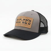 A Buc-ee’s “A Southern Tradition” trucker hat with gray front panel, black mesh back, black curved brim, and leather patch with Buc-ee’s logo, displayed on a clean white background.