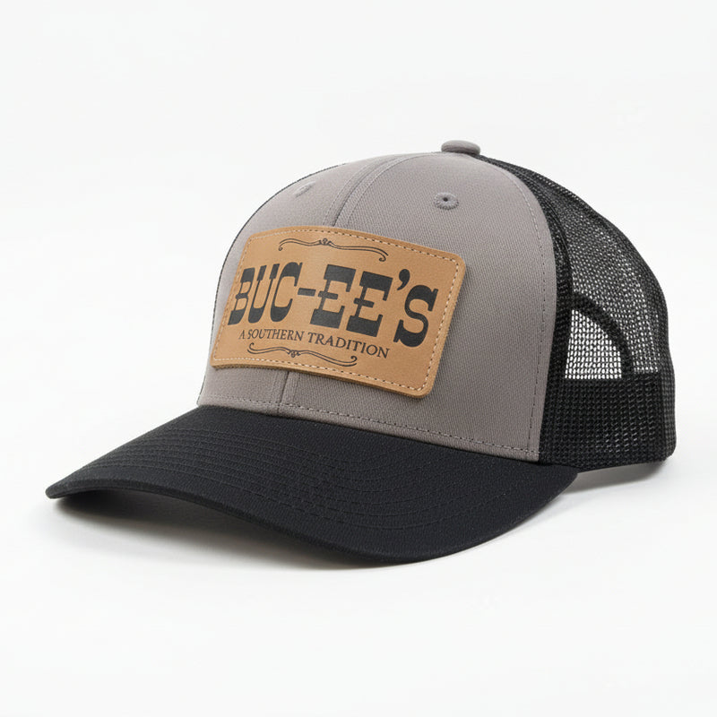 A Buc-ee’s “A Southern Tradition” trucker hat with gray front panel, black mesh back, black curved brim, and leather patch with Buc-ee’s logo, displayed on a clean white background.