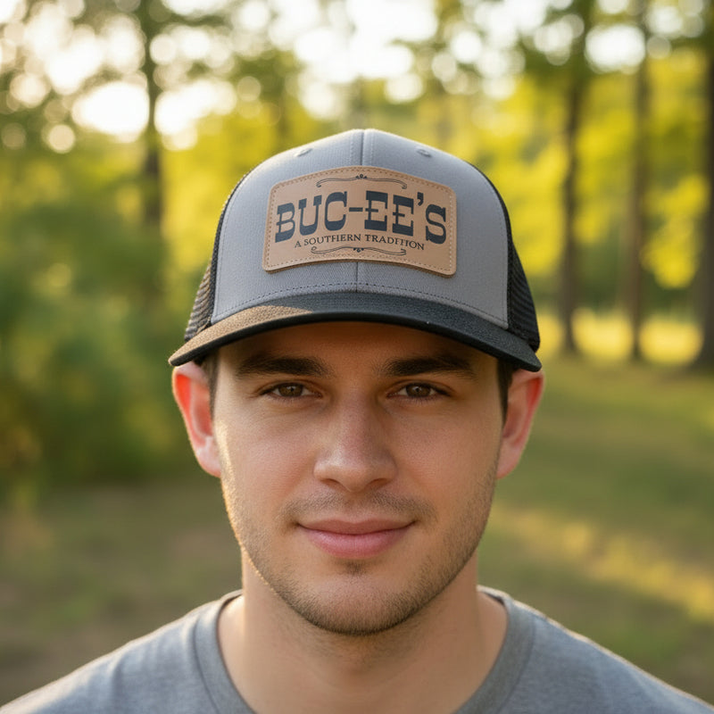A front view of a man wearing the Buc-ee's "A Southern Tradition" Trucker Hat. Featuring a gray front, black mesh sides, black curved brim and leather patch with Buc-ee's logo.