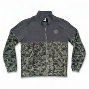 A front flat-view photo of a Buc-ee's men's full-zip fleece jacket on a white background. The jacket features a solid charcoal grey upper section, a dark camouflage lower body and sleeves, a round grey Buc-ee's beaver logo patch on the left chest, and side zippered pockets.