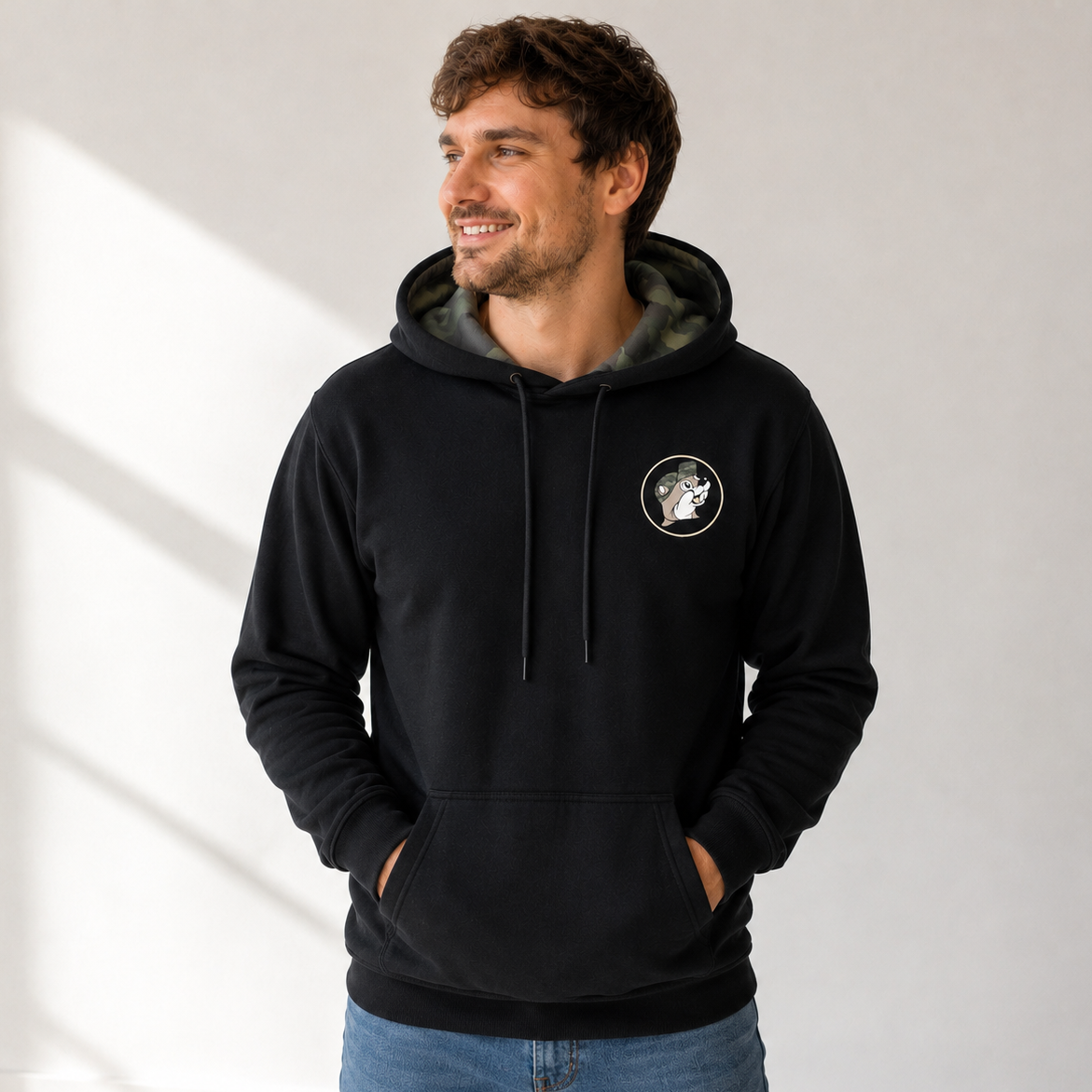 Man wearing a black Buc-ee’s hoodie with a camouflage-lined hood, drawstrings, circular beaver logo on the chest, and kangaroo pocket, paired with blue jeans.