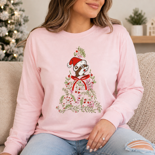Close-up lifestyle image of an adult wearing a light pink long-sleeve Buc-ee’s Christmas shirt featuring a beaver in a Santa hat surrounded by greenery, berries, and a red bow, with a cozy holiday background.
