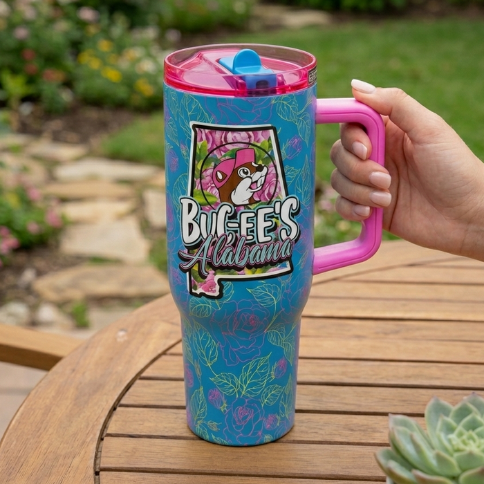 A product-focused lifestyle photo of the 40 oz Buc-ee's Alabama Fit Forty Pro Tumbler sitting directly on a wooden slatted outdoor table. A person's hand is holding the large pink ergonomic handle on the right. The teal-blue tumbler features a prominent graphic of the Alabama state outline filled with pink roses, the Buc-ee's beaver mascot, and the text "Buc-ee's Alabama." The translucent pink lid features a bright blue flip-top tab. The background shows a soft-focus, lush backyard garden with green grass
