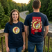 A lifestyle photo of a woman and man wearing matching navy blue Buc-ee's Alabama t-shirts on a wooden overlook. The woman faces forward, showing the front of the shirt with its pale yellow chest logo. The man has his back turned, displaying the large back graphic featuring a red Alabama state outline, a pale yellow circle, and the text "Ala-Buc-ee-Bama."