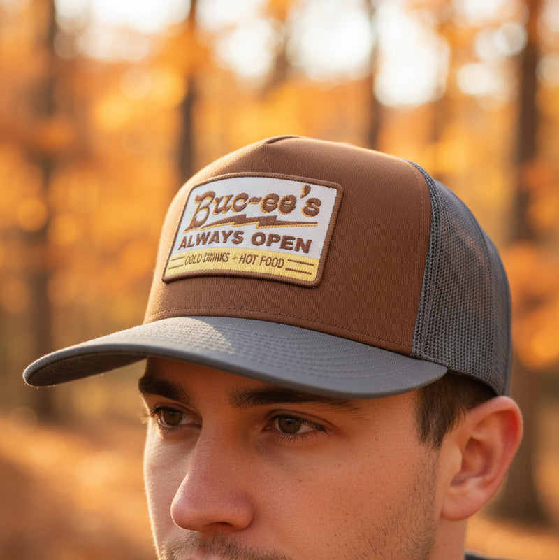 Front view of a brown and gray Buc-ee’s trucker hat featuring a rectangular embroidered patch that reads “Buc-ee’s Always Open – Cold Drinks • Hot Food,” displayed on a wooden floor