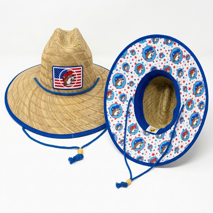 A product photograph of two Buc-ee's straw lifeguard hats against a plain white background. One hat is shown from the front, featuring a woven straw texture and a rectangular patch with an American flag background and the Buc-ee’s beaver mascot. The second hat is tilted to show the underside of the wide brim, which is lined with a vibrant blue and white fabric featuring a repeating pattern of the Buc-ee’s mascot and red and blue stars. Both hats include a blue adjustable chin cord with a wooden bead.