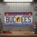 A lifestyle photograph of an Arizona Buc-ee's collectible license plate hanging on a metal pegboard wall above a wooden garage workbench. The purple and blue license plate features the word "ARIZONA" across the top, "BUC-EE'S" in large dark letters across the center with the chain's beaver mascot logo inside the letter "O," and "Beaver Believer" at the bottom. The license plate is the central focus, illuminated by a fluorescent light above. The workbench below features scattered tools.