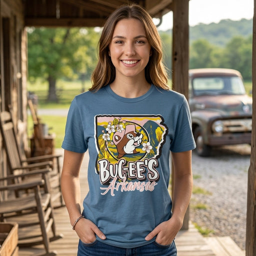 A woman stands on a rustic wooden porch wearing a blue graphic T-shirt with a cartoon bug-eel design and the words 'Bug-Eel's Arkansas.' The background shows outdoor scenery with trees, a porch, rocking chairs, and a vintage car.