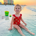 A happy toddler girl sits in the shallow, turquoise ocean water on a white sand beach at sunset. She is wearing a Buc-ee's Red Logo one-piece swimsuit with ruffle details on the shoulder straps. The swimsuit is covered in a dense, repeating pattern of the yellow and red Buc-ee’s beaver logo. In the background, there is a blurred colorful beach bucket, a small matching yellow toy shovel, a few scattered seashells, and a large, colorful beach resort building under a warm, golden-orange sky.