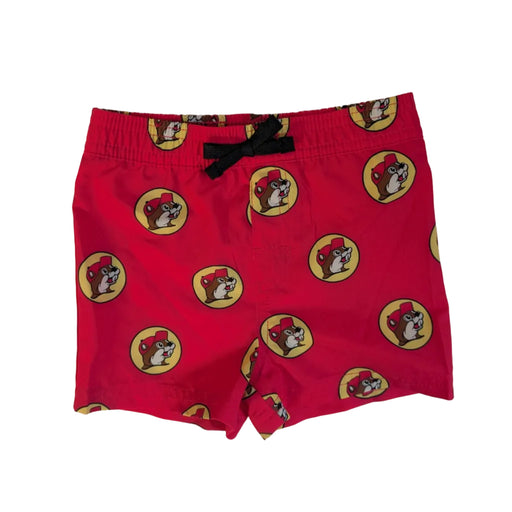 A flat lay view of bright red swim shorts featuring an all-over repeat pattern of the circular yellow and red Buc-ee’s beaver logo. The shorts include a black drawstring at the elastic waistband and are laid flat against a neutral background to show the design and fit.