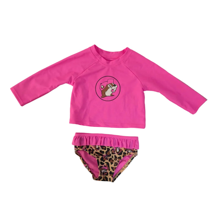 A two-piece girls' swim set featuring a solid bright pink long-sleeve rash guard top with a large circular Buc-ee's beaver mascot centered on the chest. The matching bikini bottoms feature a leopard print pattern with pink Buc-ee's icons and a pink ruffled waistband, displayed on a white background.