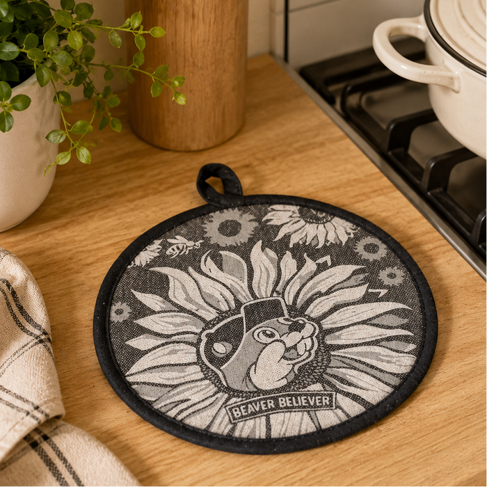 Buc-ee’s Beaver Believer heat-resistant hot pad resting on a wooden kitchen countertop beside a stove, featuring a sunflower-inspired beaver design in gray and white with black trim, displayed in a cozy kitchen setting with neutral décor and natural lighting.