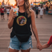 A woman stands on a crowded beach boardwalk at sunset, holding a child's hand and a colorful snow cone. She is wearing a black Buc-ee’s bling tank featuring a shimmering rhinestone beaver logo and light-wash denim shorts. A solid black fanny pack is worn around her waist. In the background, a large Ferris wheel, palm trees, and the warm glow of the setting sun create a festive summer atmosphere.