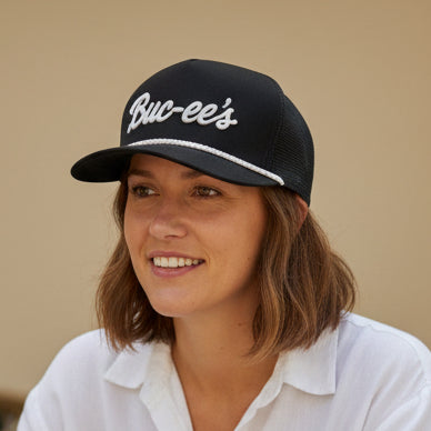 A woman wearing a Black Buc-ee’s hat with white embroidered logo and white rope detail along the brim, shown from the front.