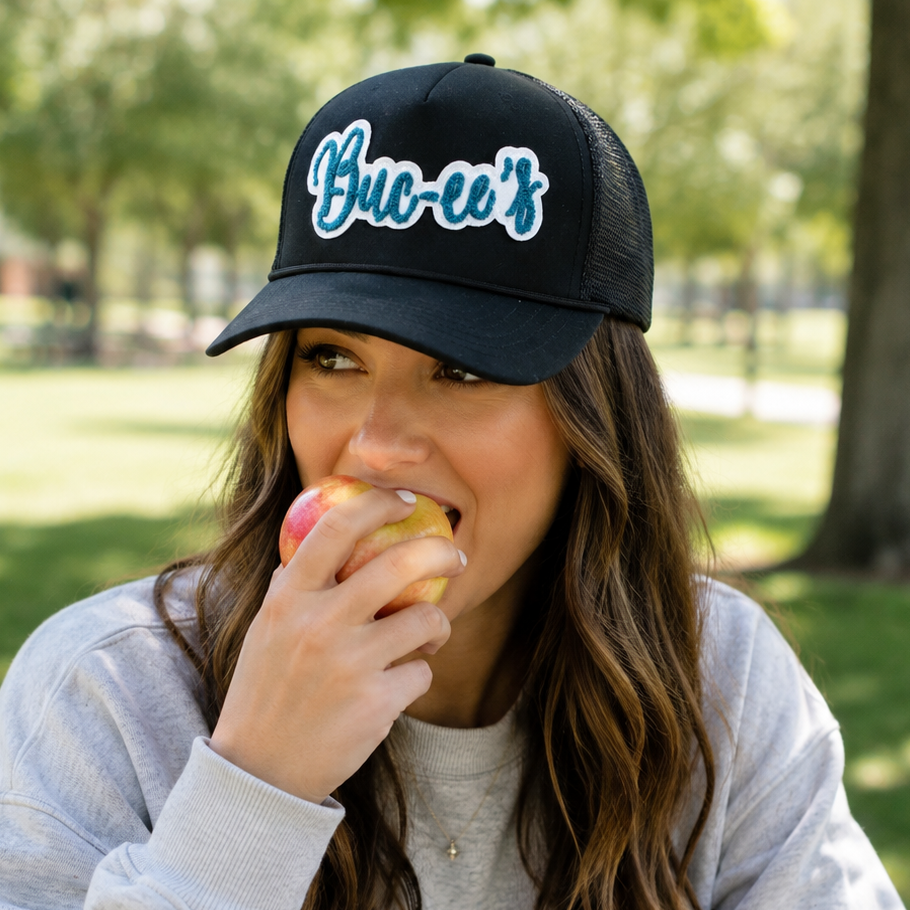 Buc-ee’s Black Trucker Hat with Teal Script Logo on a woman eating an apple.