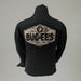Back view of a black long sleeve Buc-ee’s shirt featuring a large camouflage patch with the Buc-ee’s logo and “BUC-EE’S” text, displayed on a mannequin in front of a white draped background.
