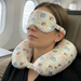 A female passenger sleeps in her plane seat wearing the Buc-ee's Blossoms and Bows Neck PIllow and Eye Mask Set, a light green base, matching pink ribbon, blue floral, and Buc-ee the Beaver logo patterns on the neck pillow and eye mask. She is in a black shirt and sits in a window seat, with clouds and a plane wing visible. Across the aisle, a male passenger in a blue shirt sits in a gray seat, and in the background, another man is visible. 