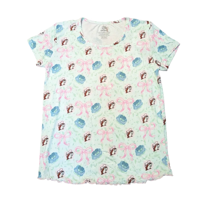 A top-down view of the Adult Blossoms & Bows Short Sleeve Pajama Shirt. The shirt is a very light green with a repeating pattern of pink bows, blue flowers, and the Buc-ee's beaver mascot wearing a matching hat. It features a classic crew neckline and a ruffled, pink lettuce-edge hem at the bottom. Displayed on a clean white background.