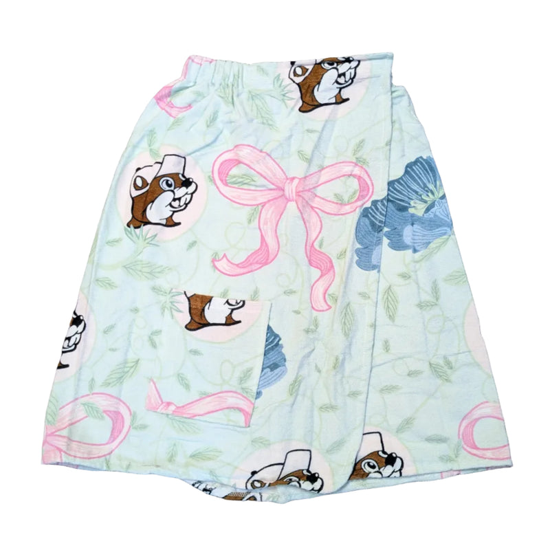 A Buc-ee’s Blossoms and Bows towel wrap laid flat against a white background. The wrap features a light mint green and white leaf pattern decorated with large pink bows, blue flowers, and circular portraits of the Buc-ee’s beaver mascot. It includes an elasticized top waistband and a matching patterned front pocket on the left side.