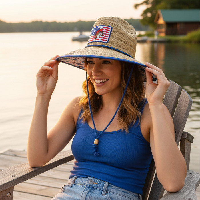 A woman with wavy brown hair sits in a weathered wooden Adirondack chair on a dock by a tranquil lake at sunset. She is wearing a royal blue tank top and denim shorts. She is holding the brim of a Buc-ee's lifeguard hat, which reveals a vibrant underside pattern of repeating Buc-ee’s beaver mascots, red stars, and blue stars on a white background. The front of the hat features a straw weave with a patch showing a Buc-ee's beaver logo over an American flag design, and it has a blue chin cord.