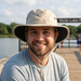 A lifestyle photo of a smiling man with a beard outdoors on a wooden fishing pier. He is wearing the Buc-ee’s Khaki Boonie Bucket Hat, which features a wide brim with a black underside and a khaki-colored trim. The hat has the signature circular beaver logo on the crown and a khaki chin strap with an adjustable toggle. The background shows a calm lake and distant trees under a bright, sunny sky, capturing a relaxed moment of outdoor recreation.