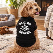 Curly-haired dog in a cozy living room wearing a black "Buc-ee the Beaver 1982" pet shirt, back view