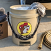 A lifestyle photo of the tan Buc-ee’s 5-gallon bucket being used for car detailing. The bucket, filled with soapy water and a grey microfiber wash mitt, sits on a concrete driveway next to a vintage tan pickup truck. Beside the bucket is a brown cardboard box labeled "Premium Detailing Solutions" topped with folded grey microfiber towels and a detailing brush. A brass-nozzle garden hose is coiled on the ground to the right. 