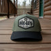 Front view of a Buc-ee’s hat with an olive-green crown, black bill, and a camo patch featuring the Buc-ee’s logo and text on the front, displayed on a cabin porch.