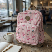 A photo of a Pink Buc-ee's Cherry Backpack with pink vertical stripes, featuring patterns of cherries, bows, and Buc-ee the beaver in a teal hat. A brown circular patch is on the upper center, and a brown base. It sits on a worn wooden table, next to a speckled ceramic mug filled with coffee. An open, lined notebook with a golden pen is to the left. The background is a brightly lit, blurred coffee shop with tables and patrons. Natural light streams in from large windows, casting soft reflections.