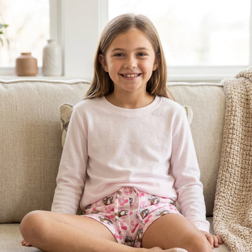 A full-body lifestyle photo of a young girl sitting happily on a beige sofa, smiling directly at the camera. She is wearing a soft, long-sleeved pink top and the light pink and white striped Buc-ee's cherry and beaver print lounge shorts. Her legs are crossed, and the shorts are clearly visible. A knit blanket is on the sofa beside her.
