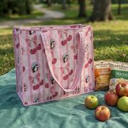 A Buc-ee's Cherry Quilted Tote Bag, a pink-and-white vertically striped tote bag with cherry and bow patterns, sitting on a teal picnic blanket with snack items in a grassy park setting. The background features blurred trees and a distant dirt path under sunny, natural light.