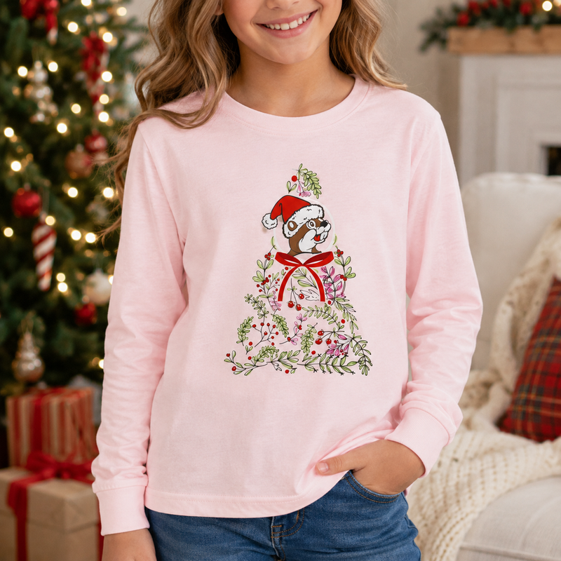 Close-up lifestyle image of a youth wearing a light pink long-sleeve Buc-ee’s Christmas shirt featuring a beaver in a Santa hat surrounded by greenery, berries, flowers, and a red bow, with a decorated Christmas tree and gifts in the background.
