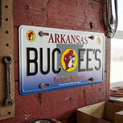 A close-up of a Buc-ee's Arkansas "Beaver Believer" metal collectible license plate mounted on a rustic wooden garage wall next to a pegboard and tools.