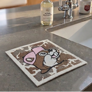 A lifestyle photograph from a kitchen island view, featuring a Buc-ee's Swedish dishcloth with cowhide patterns and smiling Buc-ee beaver face wearing a pink hat resting on a grey quartz countertop near a sink with a stainless steel faucet.