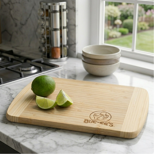 Close-up of a Buc-ee's Cutting Board, a rectangular multi-tone bamboo cutting board featuring light-colored vertical bands and a Buc-ee’s logo in the corner. The board is sitting on a marble countertop next to a stovetop and a stack of ceramic bowls. Also a lime and two slices of lime are on top of the cutting board.