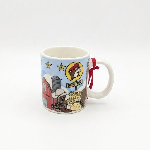 A white ceramic mini mug against a clean white background featuring a light blue sky wrapped in Texas-themed illustrations. The graphics include a red barn, a Texas longhorn steer, a taco, roses, a horseshoe, a Texas flag map outline, and a yellow Buc-ee's sign reading "DENTON". A small red hanging string is tied to the handle.