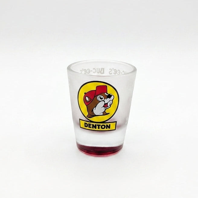 A tapered, frosted clear glass shot glass featuring the iconic yellow circular Buc-ee's beaver logo with a yellow banner reading "DENTON" underneath. The heavy, solid glass base is tinted in a vibrant, translucent red color.