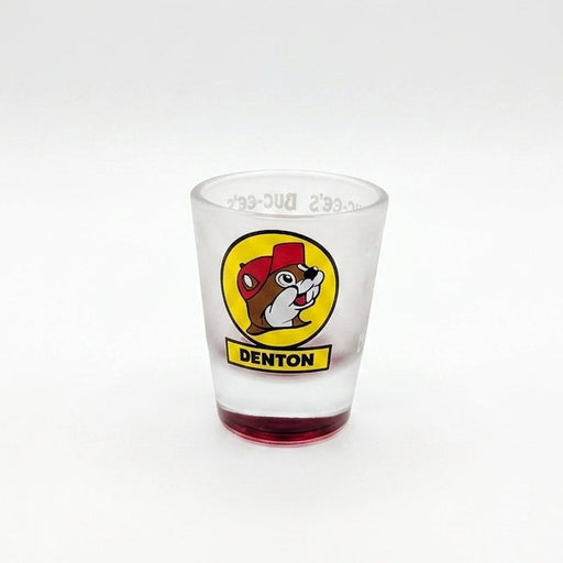 A tapered, frosted clear glass shot glass featuring the iconic yellow circular Buc-ee's beaver logo with a yellow banner reading "DENTON" underneath. The heavy, solid glass base is tinted in a vibrant, translucent red color.
