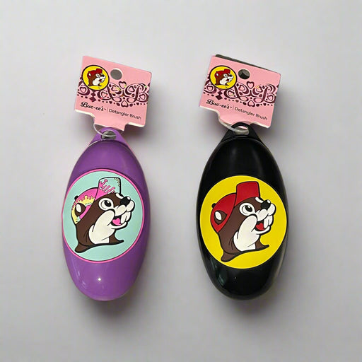 Buc-ee’s detangler brushes in two colors: one black with Buc-ee in a red cap, and one purple with Buc-ee surrounded by pink hearts, shown from front and back views.

