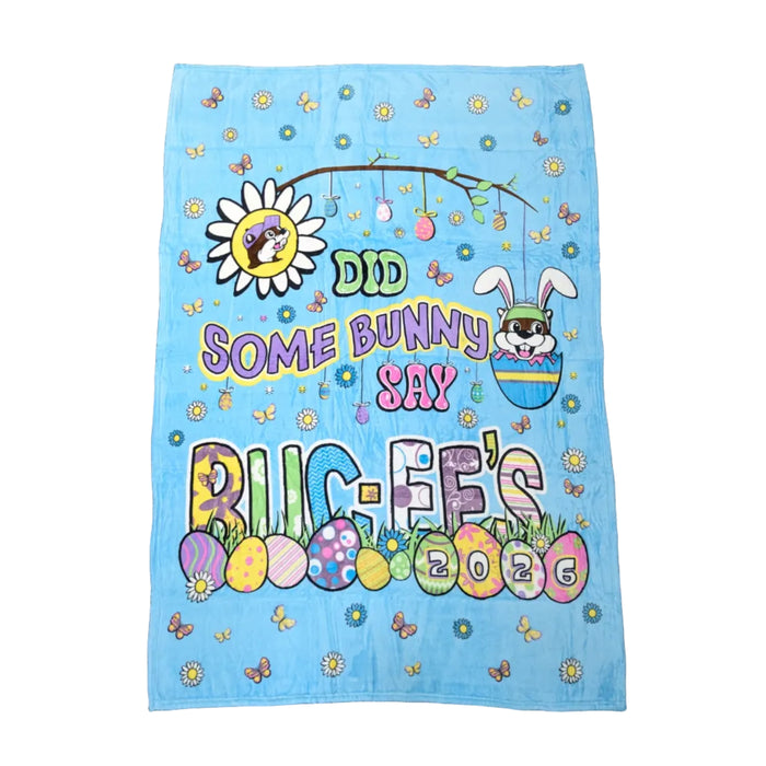 A large, rectangular light blue plush blanket laid flat. The center features the text "DID SOME BUNNY SAY BUC-EE'S" in colorful bubble letters. The design includes Buc-ee the beaver wearing bunny ears inside a blue egg, a daisy with Buc-ee's face in the center, and various decorated Easter eggs along the bottom, one of which displays the year "2026".