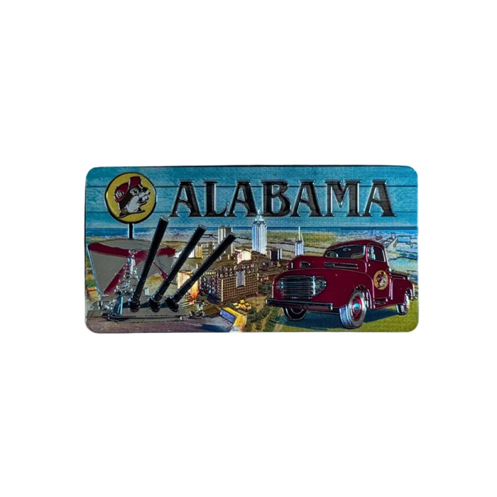 One side of a rectangular metallic Buc-ee's Double-Sided Alabama souvenir magnet laid flat on a white surface. This side of the magnet features a scenic illustration under a teal sky featuring the word "ALABAMA," a vintage red truck, and a Buc-ee's billboard overlooking a harbor city.