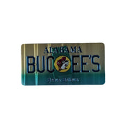 One side of a rectangular metallic Buc-ee's Alabama Double-Sided souvenir magnet laid flat on a white surface. This side of the magnet features a gold-and-green gradient background with embossed text reading "ALABAMA BUC-EE'S" and "Beaver Believer." 