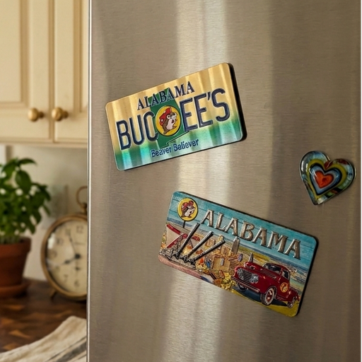 A photo of both sides of a rectangular Buc-ee's Double-Sided Alabama souvenir magnet displayed on a stainless steel refrigerator in a cozy kitchen. The top magnet features a gold and green gradient background with the text "ALABAMA BUC-EE'S" and "Beaver Believer." The bottom magnet displays a colorful scenic illustration with a vintage red truck, a harbor skyline, and a Buc-ee's billboard. A small, colorful heart-shaped magnet sits to the right. 