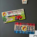 A front-facing, lifestyle photo of the Buc-ee’s Double-Sided Ohio Magnet displayed on a black magnetic board inside a light wood frame. The top magnet features a scenic Ohio landscape with a red vintage truck, a barn, and the state flag. The bottom magnet has a red, silver, and blue gradient with the "Buc-ee’s Beaver Believer" logo. Both magnets are approximately 3.5 by 1.75 inches. A small white magnet shaped like a house with the word "home" sits at the top, and a child's drawing of a person.