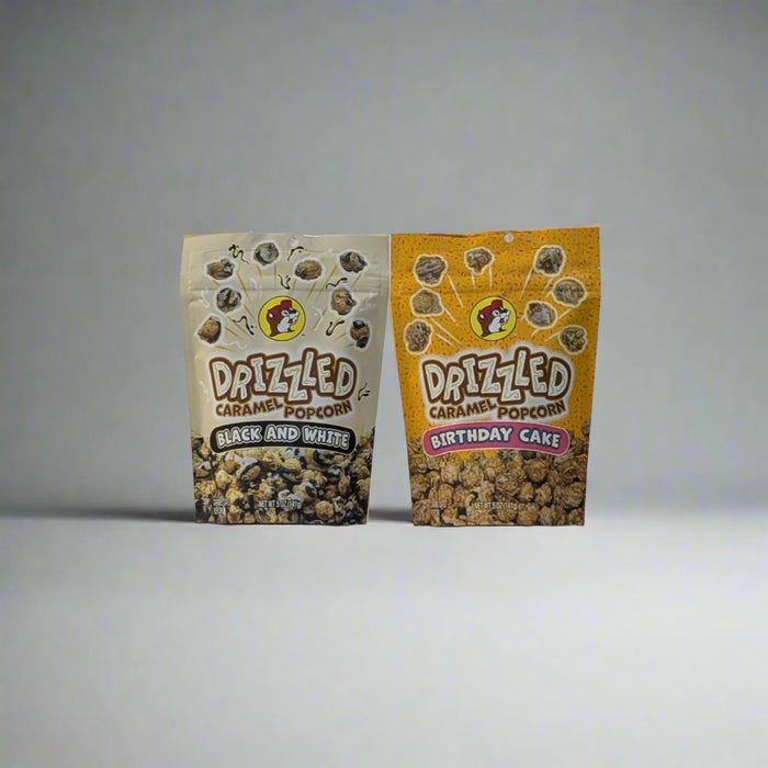Two bags of Buc-ee’s Drizzled Caramel Popcorn, one in Black and White flavor and the other in Birthday Cake flavor, displayed on a white sheet backdrop.

