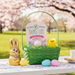 A wicker Easter basket sits on a light-colored wooden table outdoors. Inside the basket, there is a Buc-ee's 'Cotton Candy' bath bomb in its original packaging, a yellow plush chick, and green Easter grass. Outside of the basket on the table, there is a classic-style chocolate bunny and three pastel-colored plastic Easter eggs. The background shows a soft-focus, sunny spring scene with cherry blossom trees in bloom.