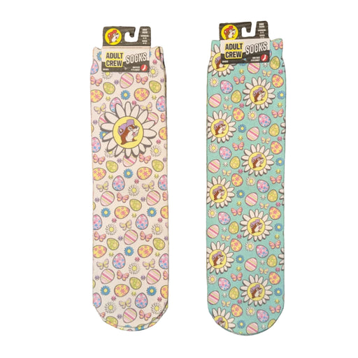 A flat-lay photo shows two pairs of Buc-ee's adult crew socks with a festive Easter theme. Both pairs feature a repeating pattern of colorful Easter eggs, butterflies, and daisies, with the beaver mascot wearing a purple hat at the center of the largest flowers. One pair has a white background, while the other has a light teal background, both are displayed on a clean white background.
