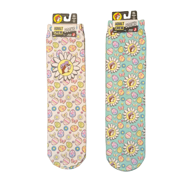 A flat-lay photo shows two pairs of Buc-ee's adult crew socks with a festive Easter theme. Both pairs feature a repeating pattern of colorful Easter eggs, butterflies, and daisies, with the beaver mascot wearing a purple hat at the center of the largest flowers. One pair has a white background, while the other has a light teal background, both are displayed on a clean white background.