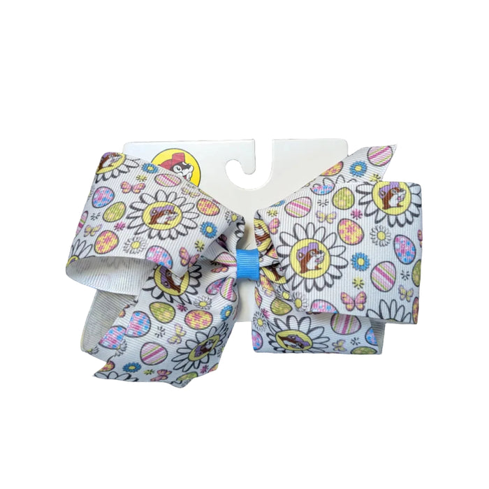 A close-up view of the Classic White Buc-ee’s Easter hair bow. The bow is made of textured ribbon with a white background and an all-over print of white daisies, pastel Easter eggs, and the Buc-ee’s beaver logo wearing a purple hat. The center of the bow is secured with a small blue fabric knot, and the retail packaging features the red-hatted Buc-ee's logo at the bottom. Displayed on a clean white background.